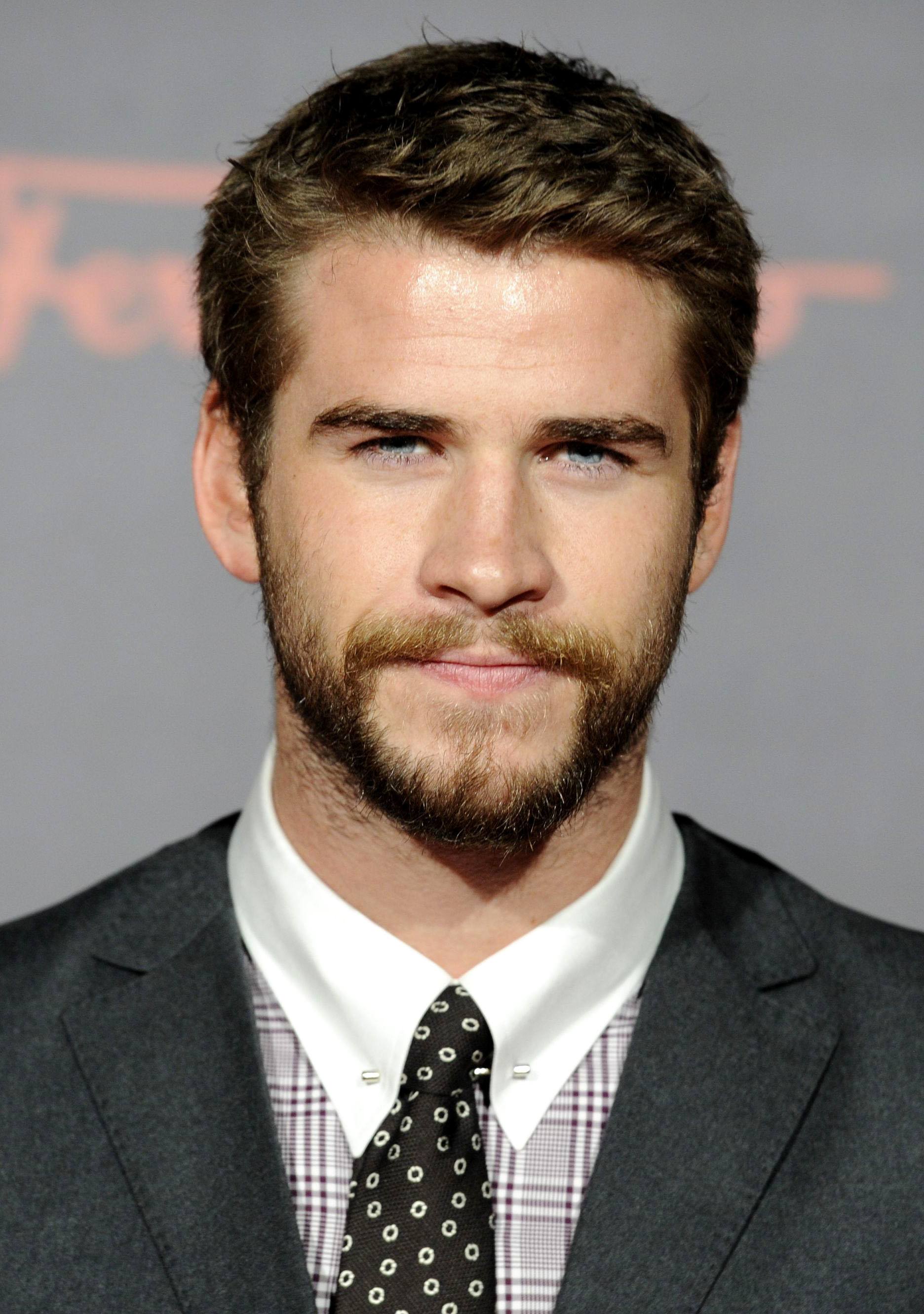 Coupe de cheveux Ivy League: Liam Hemsworth aux cheveux bruns dans une coupe de cheveux texturée Ivy League, vêtu d'un costume gris