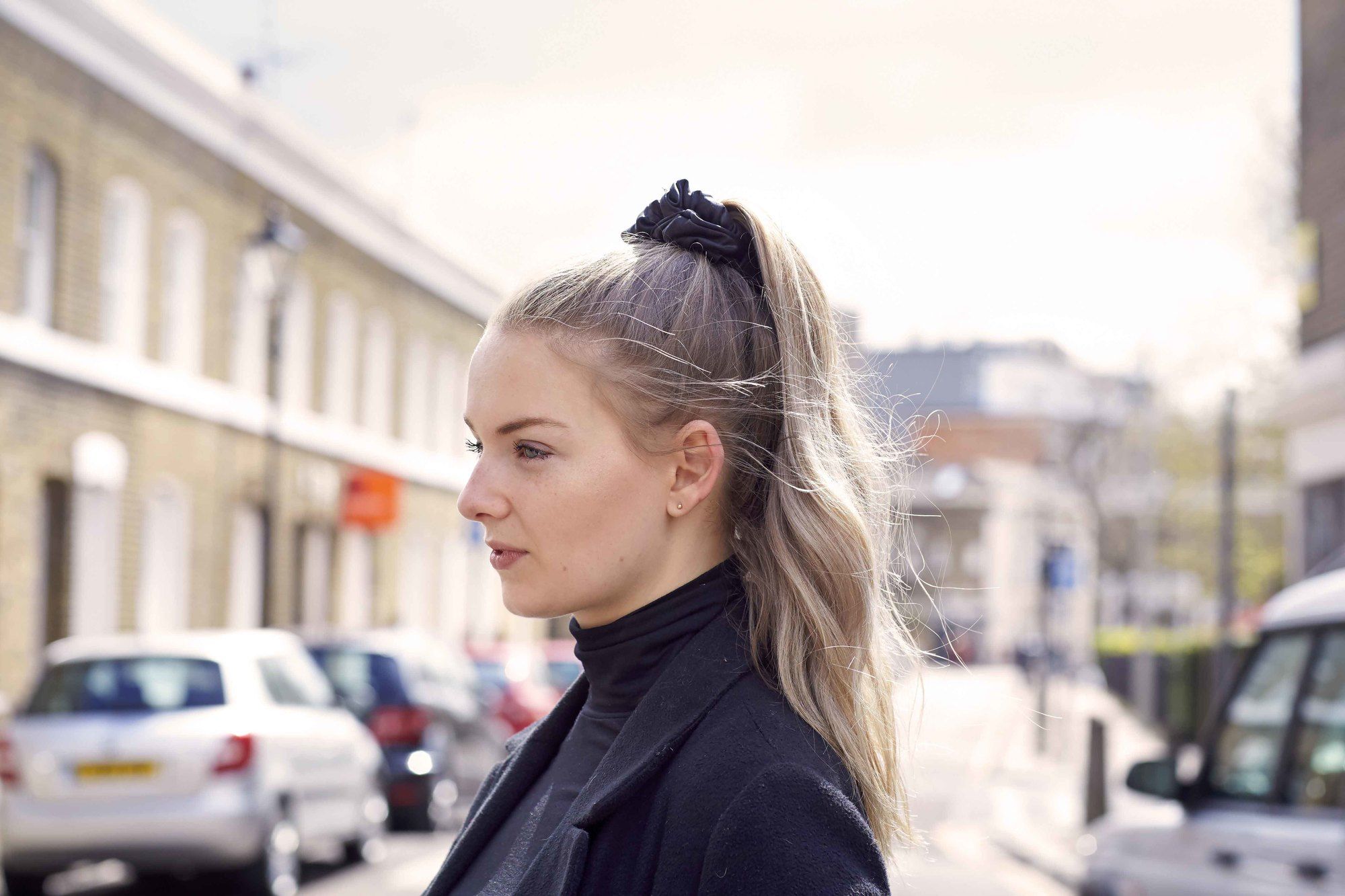 Coiffures faciles pour les cheveux longs: Femme aux cheveux ondulés surlignés blonds en queue de cheval haute avec un chouchou noir portant un haut à col roulé noir et un blazer.