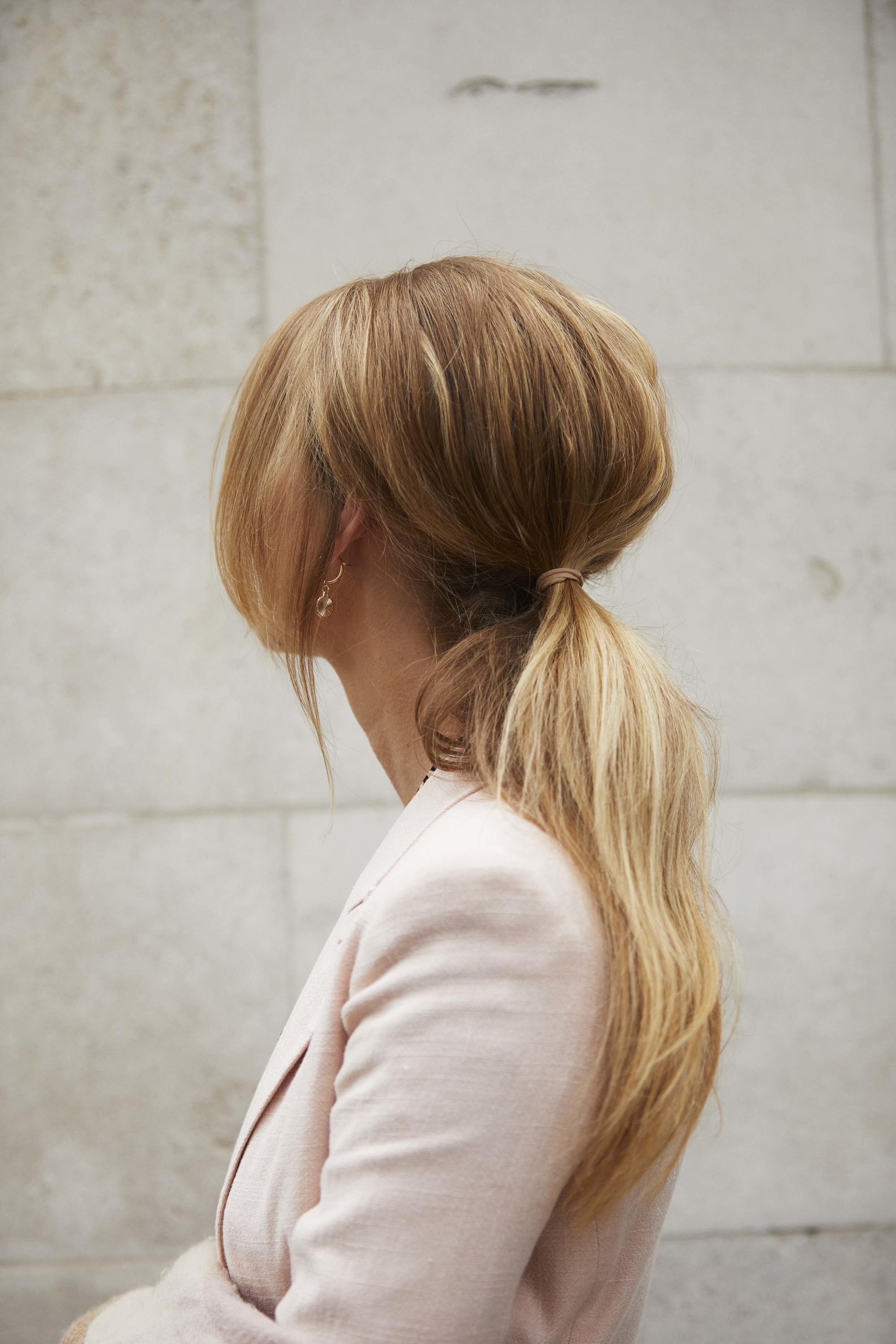 Wedding hairstyles: Woman with long blonde golden hair styled into a bouffant ponytail, wearing a pink suit jacket and posing outside