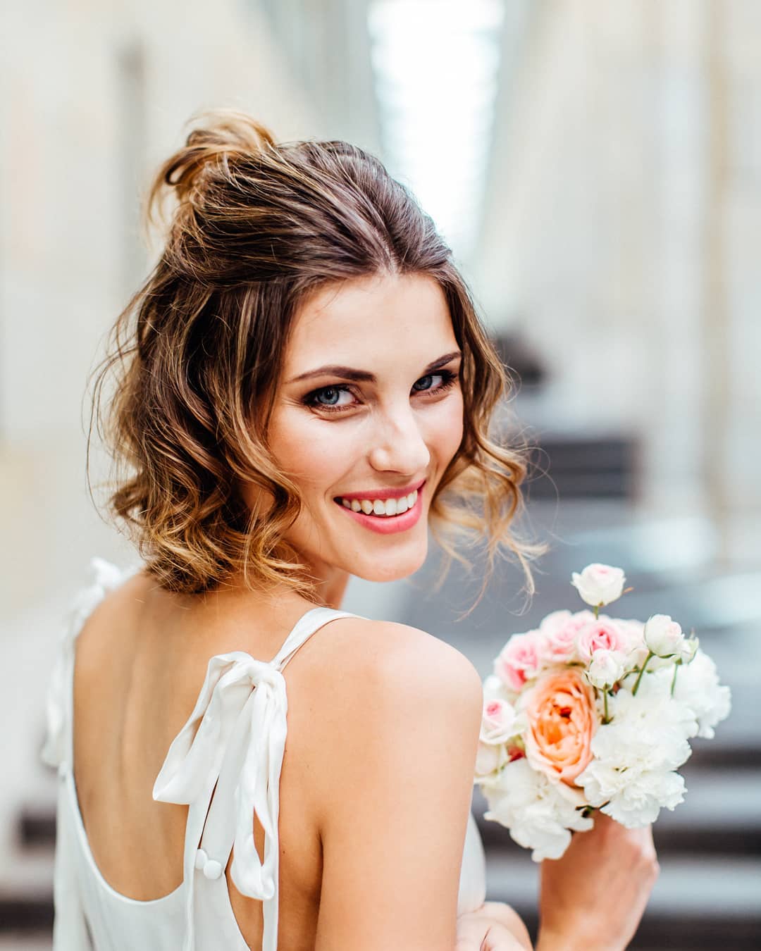 Wedding hairstyles: Woman with half-up, half-down short wavy hair, wearing a white dress and holding flowers