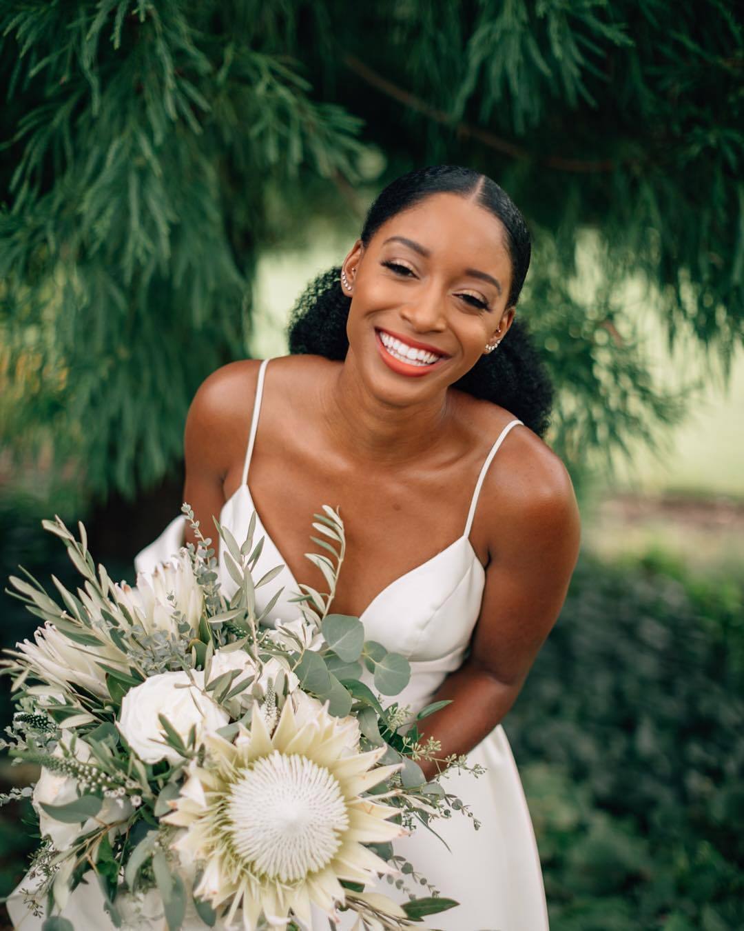 Wedding hairstyles: Woman with low, long sleek natural ponytail, wearing white dress and holding flowers