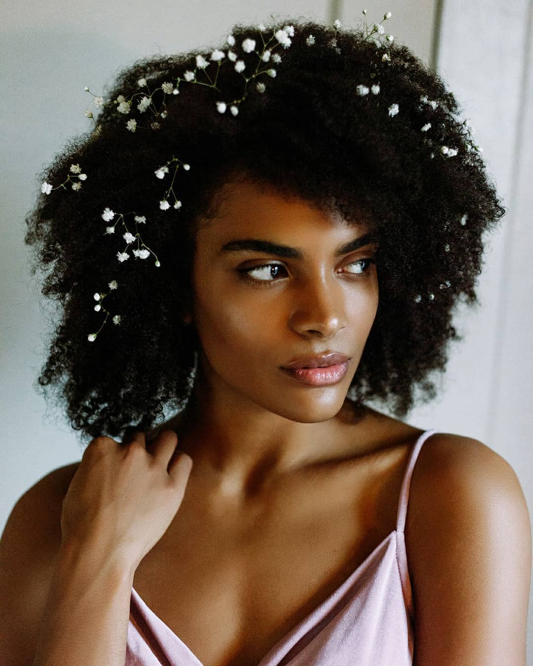 Black wedding hairstyles: Woman with dark brown afro with baby's breath flowers in it, wearing purple satin dress while posing in a bedroom setting