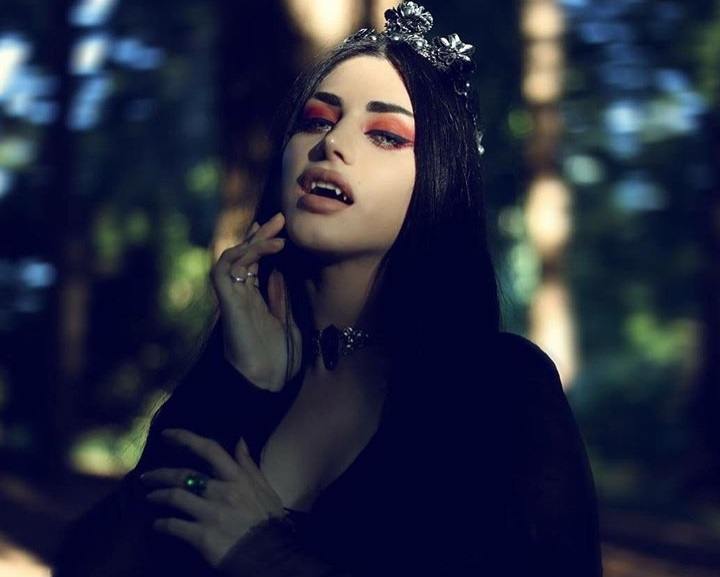 Coiffures d'Halloween : Femme habillée en vampire, avec des cheveux noirs lisses et un accessoire de coiffure en forme de diadème étincelant, portant tout en noir et posant dans un décor boisé
