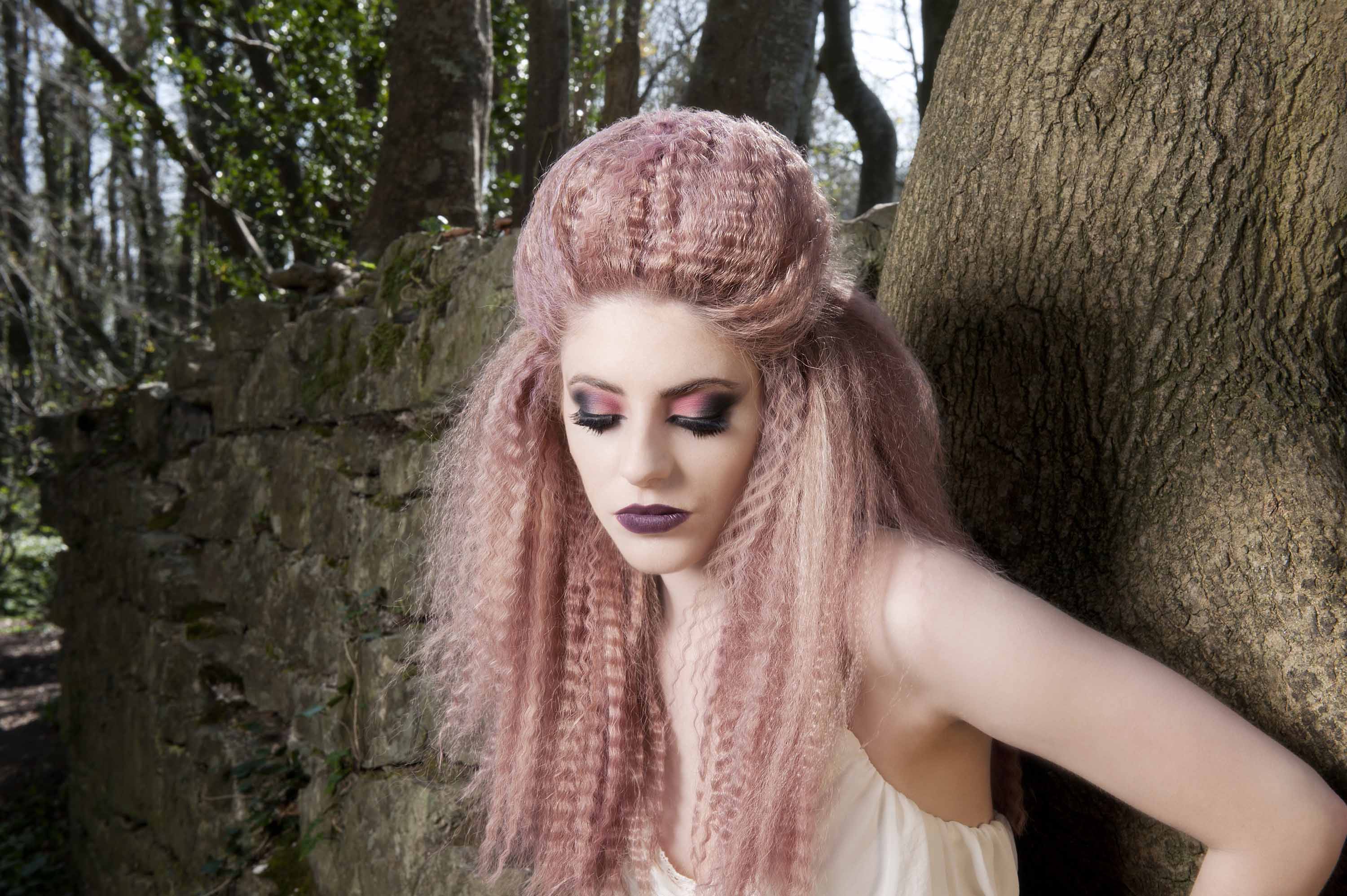 femme aux cheveux rose pastel frisés en demi-lune, appuyée contre un arbre