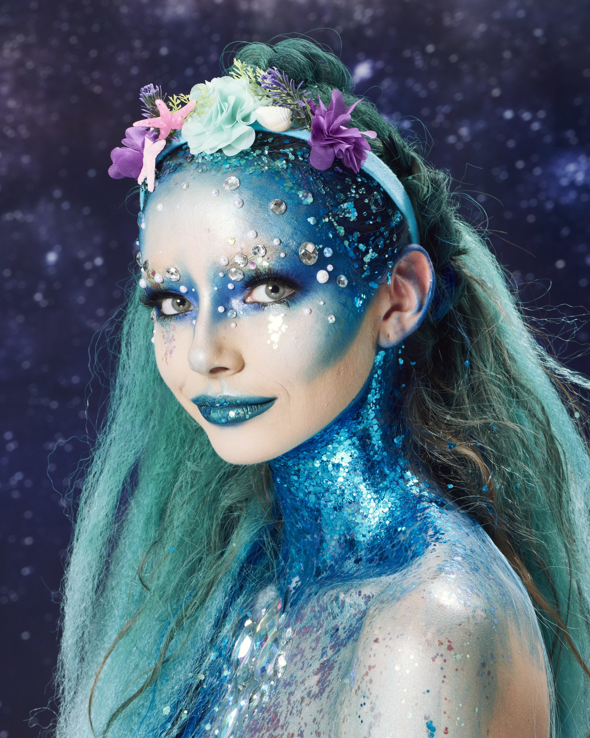 Coiffures d'Halloween : Femme habillée comme une sirène, avec de longs cheveux bleus, une tresse de licorne et un bandeau floral, portant du maquillage bleu et des paillettes bleues