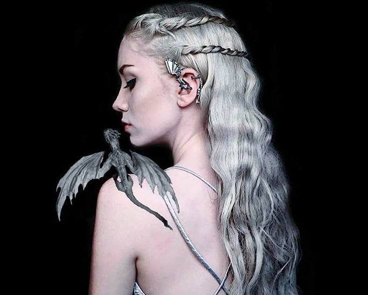 Coiffures d'Halloween : Femme habillée en Khaleesi de Game of Throne, avec ses longs cheveux blonds argentés coiffés en vagues lâches avec deux tresses, portant de l'argent et posant avec un dragon CGI sur son épaule