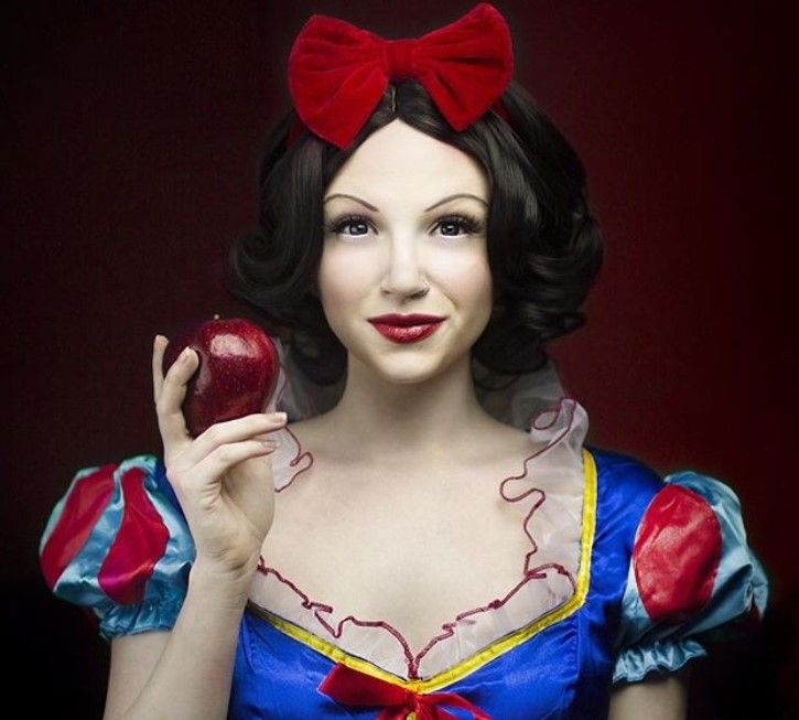 Coiffures d'Halloween : Femme habillée en Blanche-Neige, avec ses cheveux noirs courts coiffés en boucles avec un accessoire pour cheveux en noeud rouge, tenant une pomme et posant dans un décor de studio