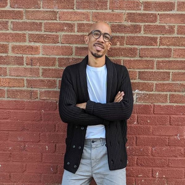Homme noir chauve à la peau claire, avec des lunettes, appuyé contre un mur de briques