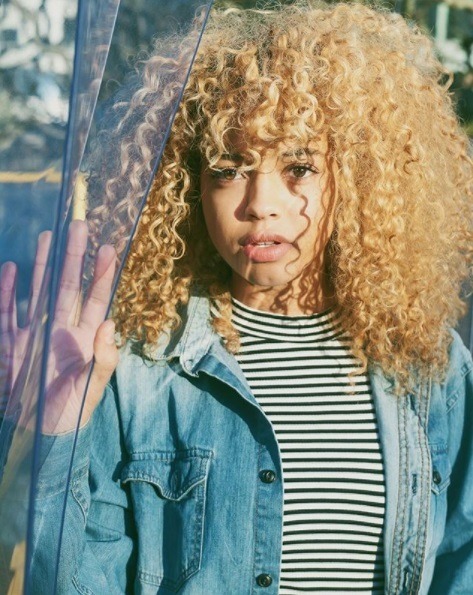 femme blonde aux cheveux naturels avec une frange pleine portant une veste en jean et un tshirt rayé