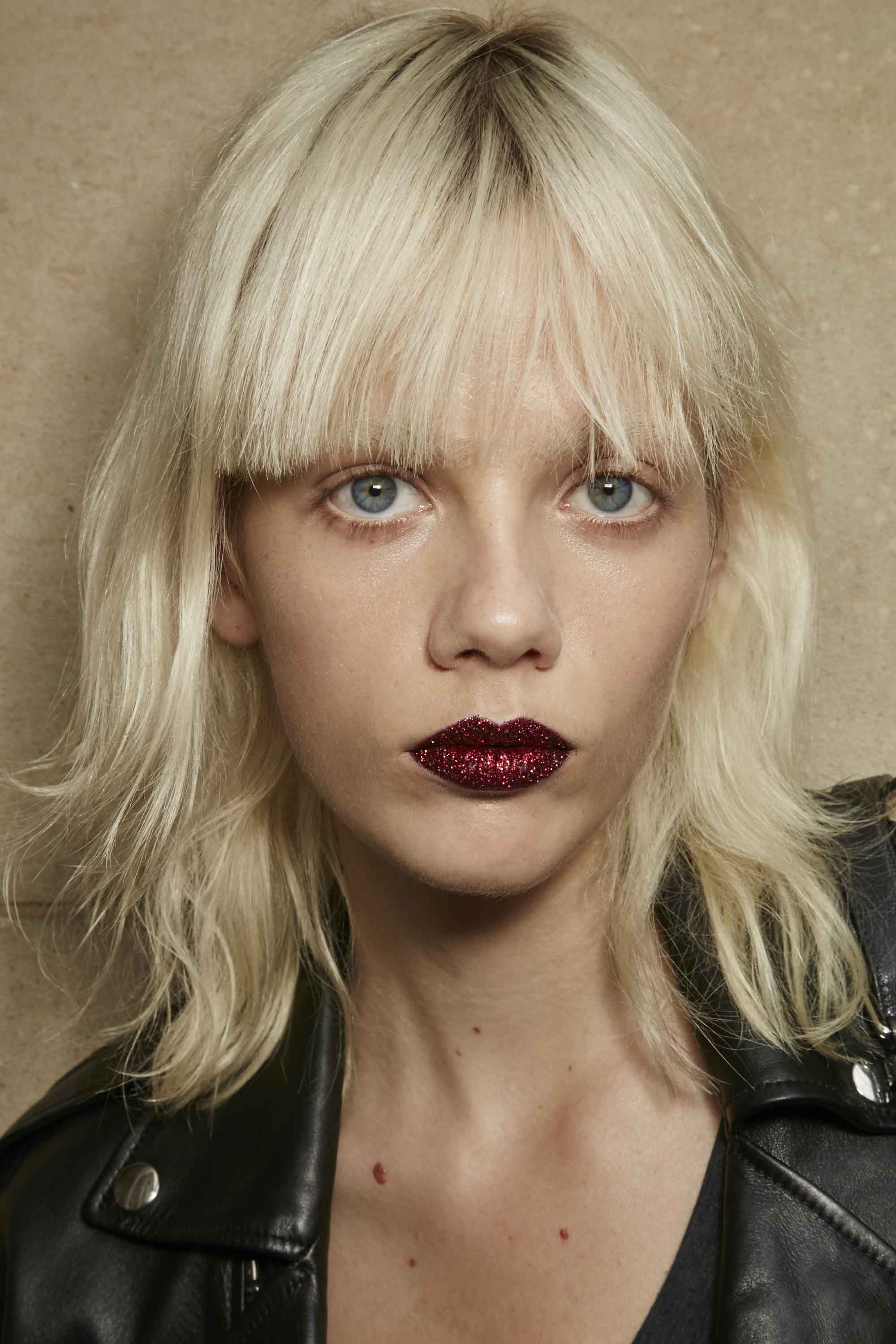 mannequin avec des cheveux blonds platine coupés en une coupe courte et une frange, portant du rouge à lèvres rouge foncé.