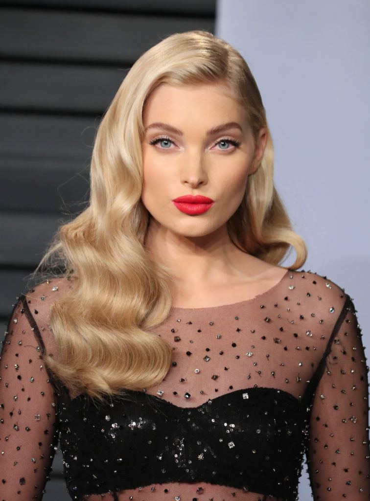 Nuances de cheveux blonds : Elsa Hosk avec une longue chevelure ondulée blonde champagne dans une raie latérale, portant du rouge à lèvres rouge et une robe noire diaphane.