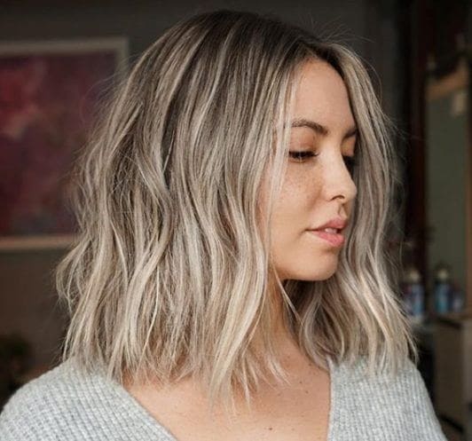 Nuances de cheveux blonds : Femme aux cheveux blonds cendrés surlignés de bob ondulé, portant un pull en tricot gris.