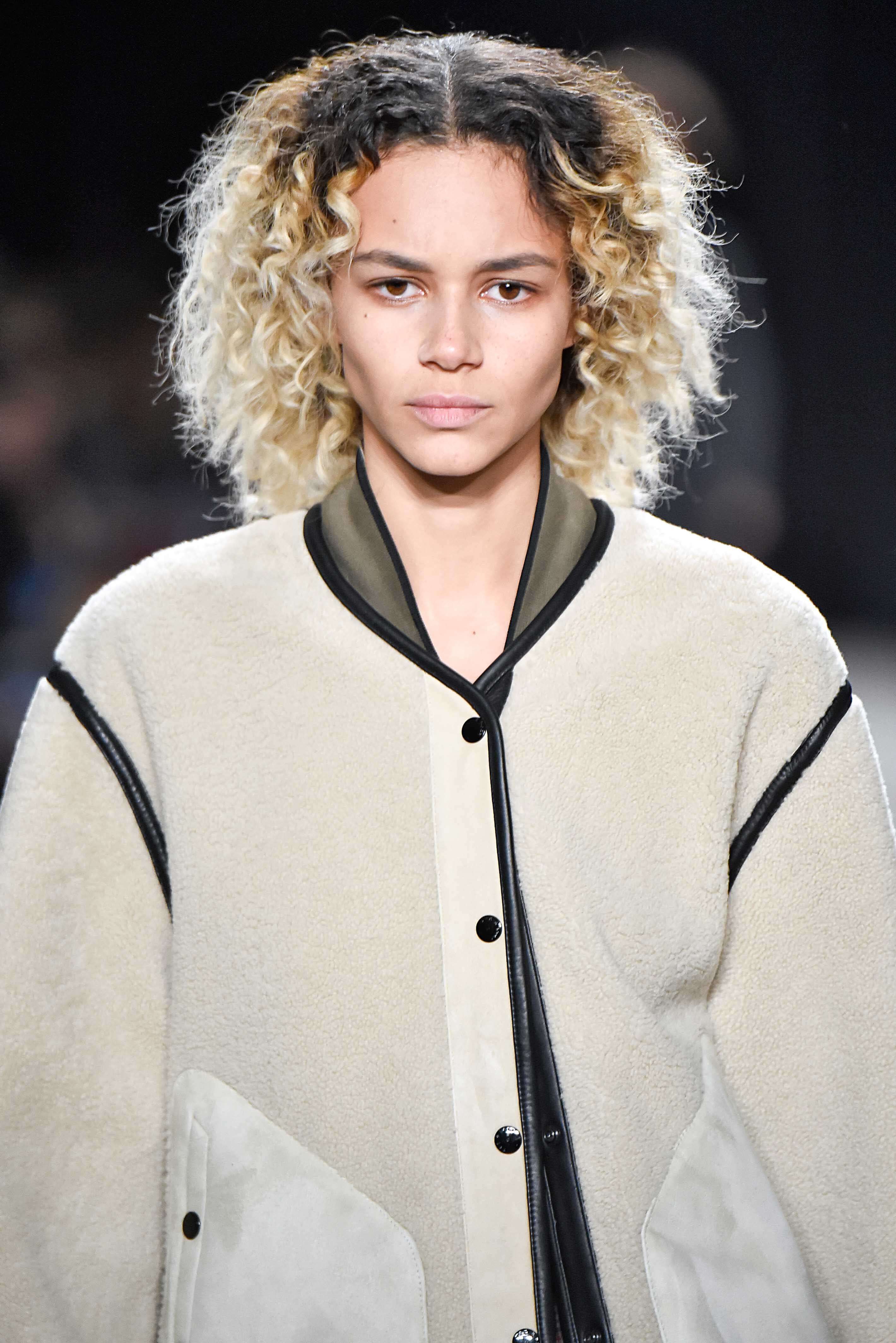 Nuances de cheveux blonds : Femme aux cheveux blond platine naturellement bouclés avec des racines brun foncé sur une piste de décollage, portant un blouson de bombardier.
