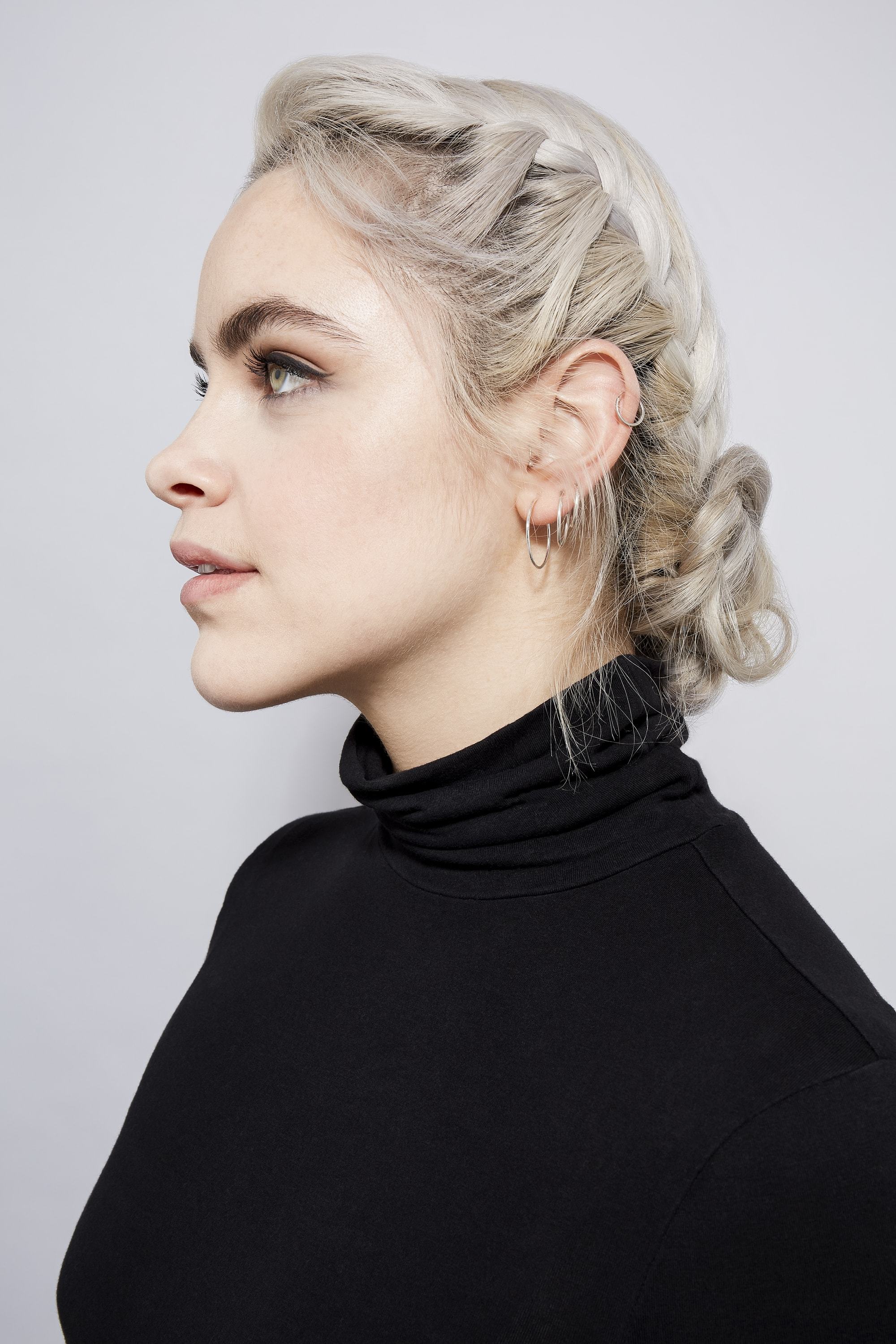 Des tresses faciles pour les cheveux longs : Profil latéral d'une femme aux cheveux blonds cendrés dans un chignon tressé, portant un top noir à col roulé
