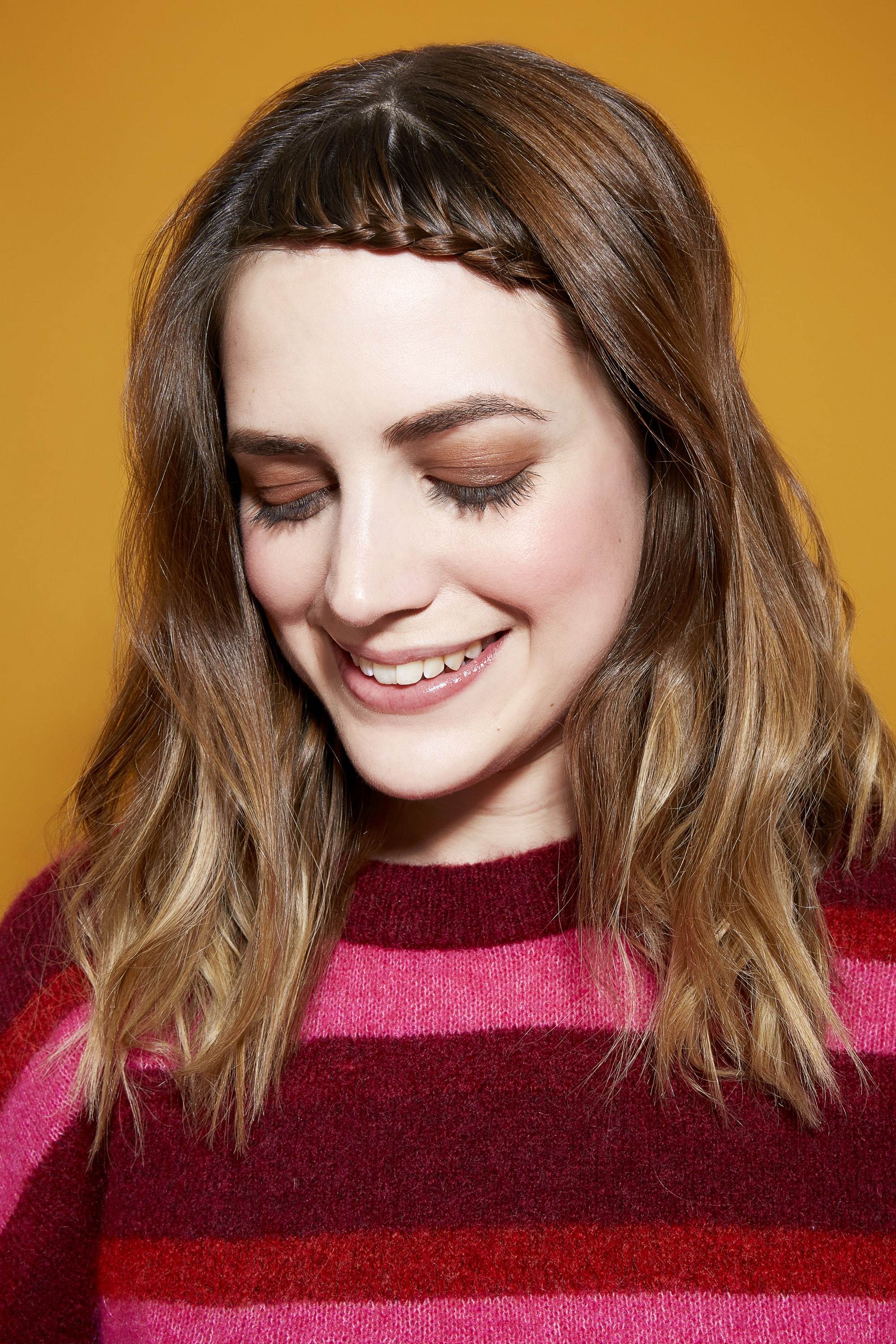 Des tresses faciles pour les cheveux longs : Femme brune à la frange tressée, portant un pull rayé rose et bordeaux