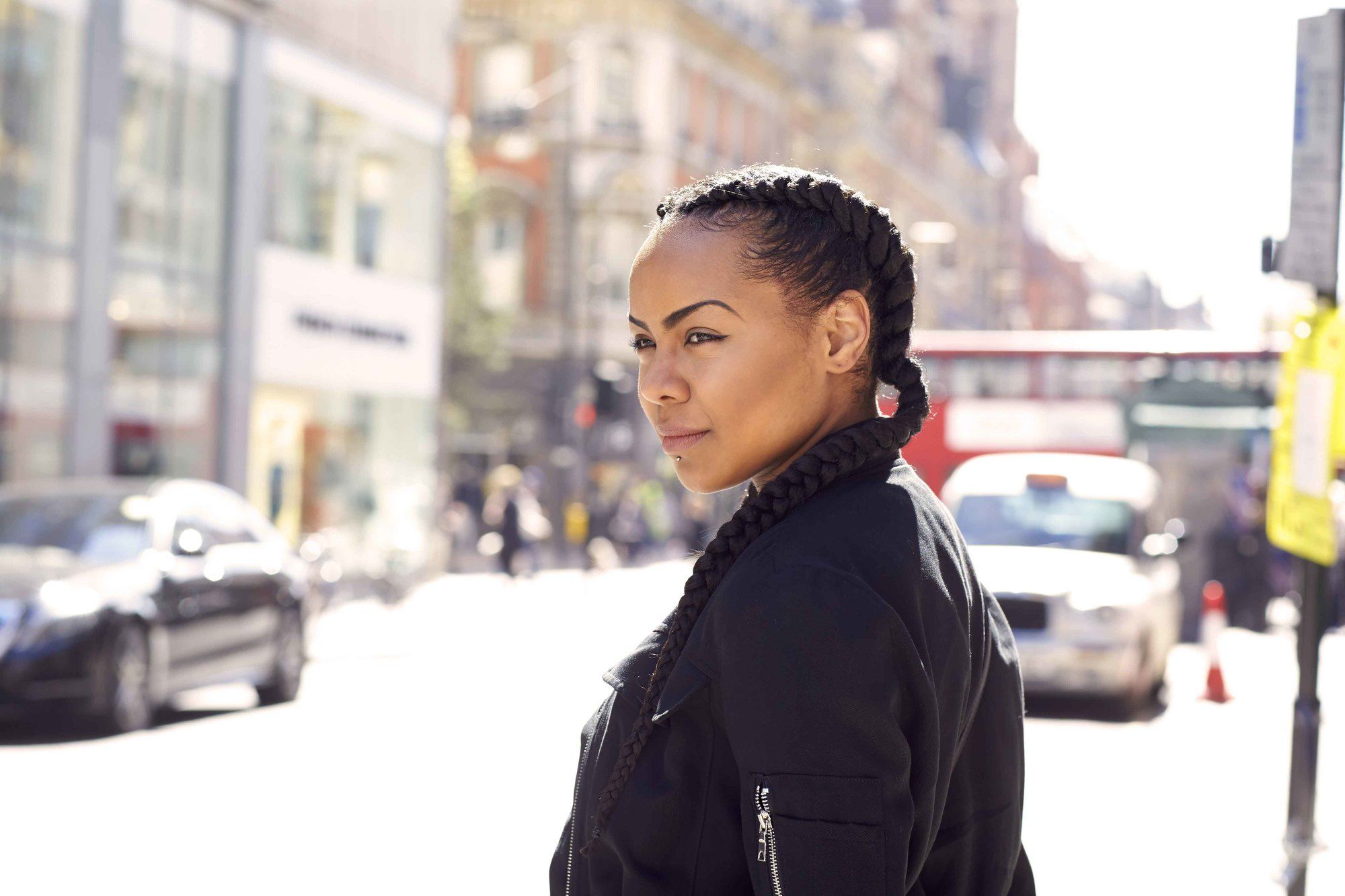 Coiffures d'hiver pour cheveux naturels : Vue latérale d'une femme avec des tresses de boxer double marron foncé, portant une veste de bombardier noire et se tenant dans la rue