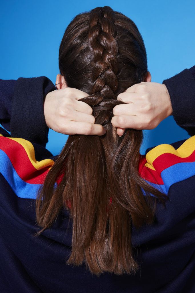 Comment faire une tresse hollandaise : Vue de dos d'une jeune fille brune tressant ses cheveux en une tresse hollandaise, portant un pull à rayures arc-en-ciel et se tenant debout sur un fond bleu