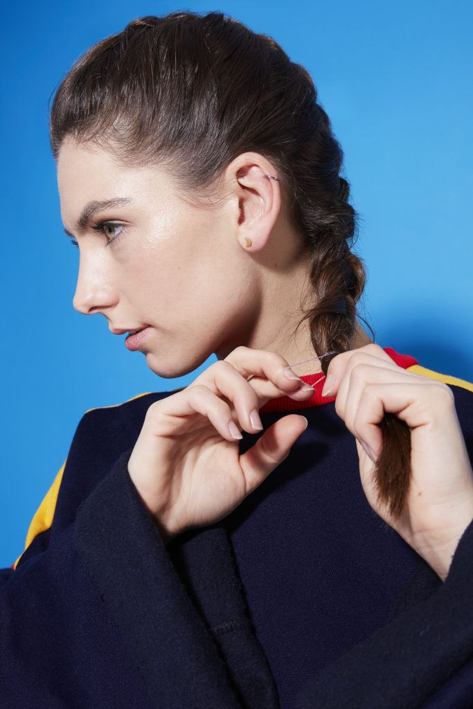 Comment faire une tresse hollandaise : Profil latéral d'une jeune fille brune nouant une tresse au bas de ses cheveux, debout sur un fond bleu