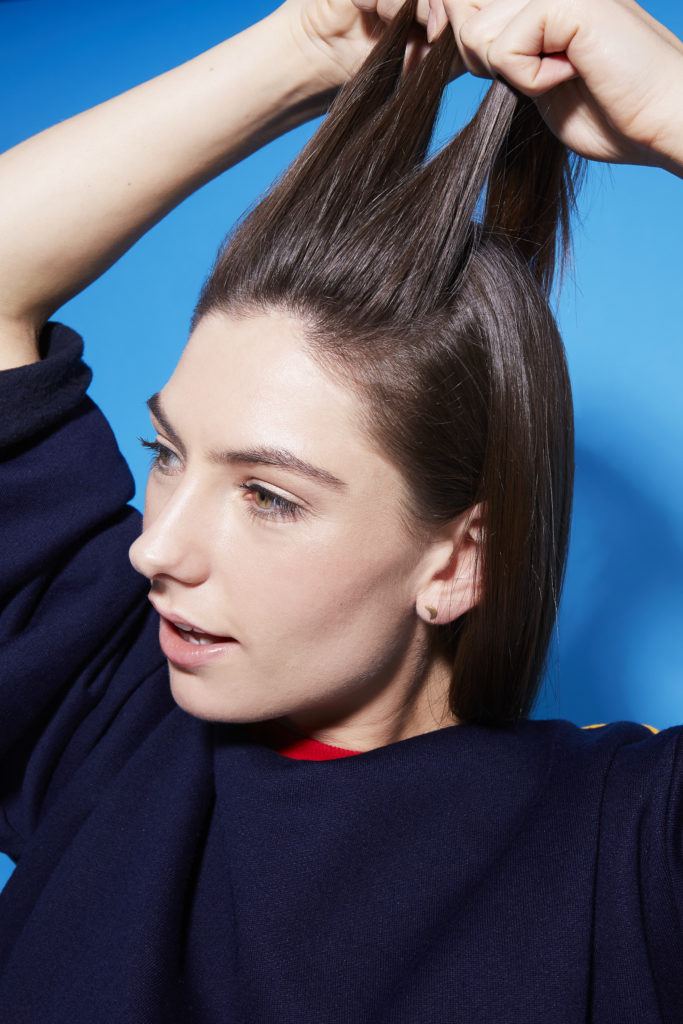 Comment faire une tresse hollandaise : Fille brune tenant des mèches de cheveux sur le devant, sur fond bleu