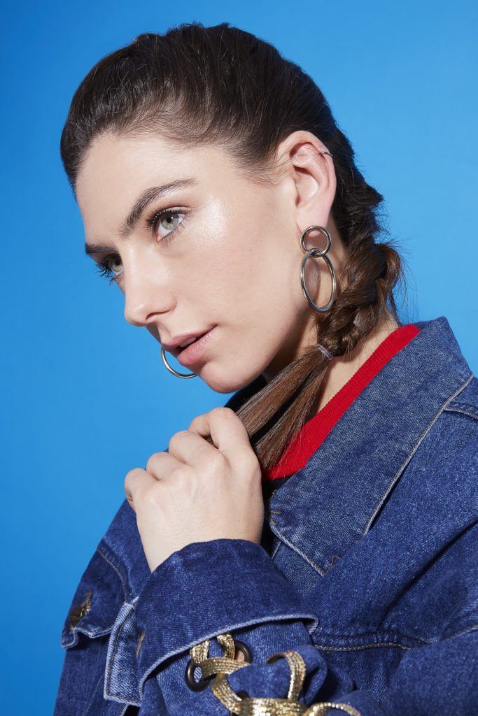 Comment faire une tresse hollandaise : Gros plan d'une jeune fille brune avec une tresse hollandaise tenant le bout de sa tresse, portant une veste en denim bleue et se tenant debout sur un fond bleu