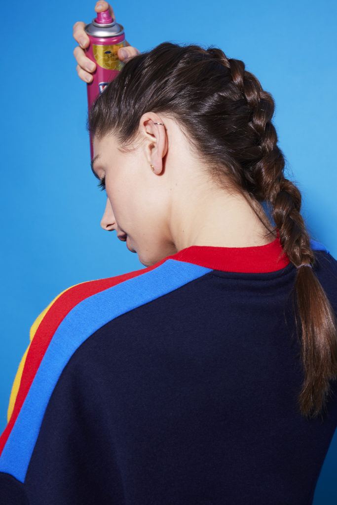 Comment faire une tresse hollandaise : Vue arrière d'une jeune fille brune avec une coiffure à tresses hollandaise qui se vaporise de la laque sur les cheveux