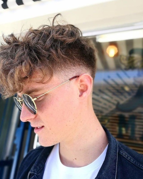 homme aux cheveux bouclés brun clair, avec un front lourd en faux mohawk dont les côtés sont décolorés