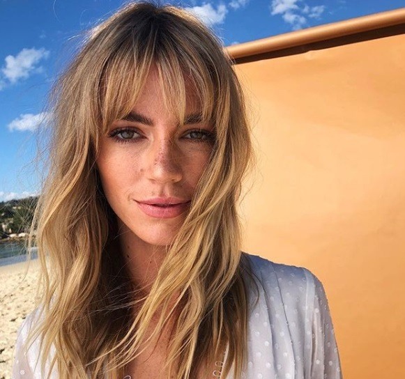 Types de franges : Femme aux longs cheveux blonds avec une pleine frange Birkin, vêtue de blanc et posant à l'extérieur.