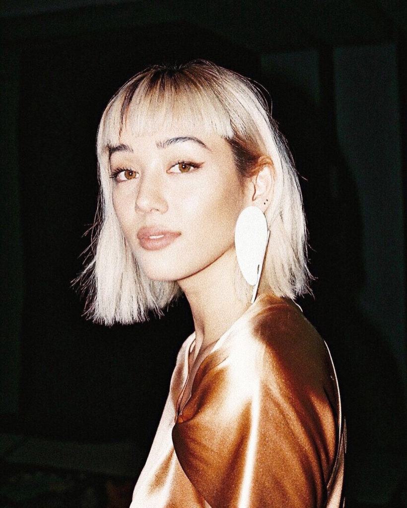 Guide des types de franges : Kim Jones avec un carré long blond décoloré coiffé d'une micro-frange, portant une boucle d'oreille en forme de coquillage avec un haut en satin orange.