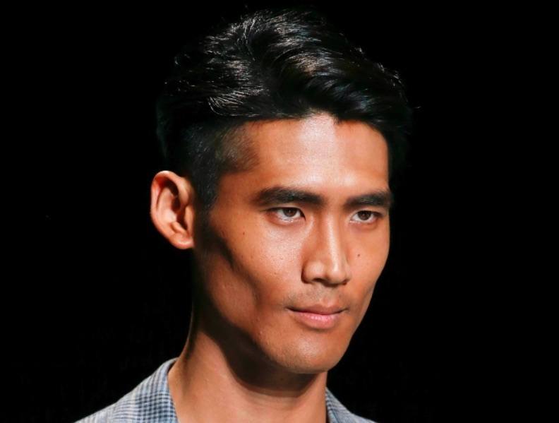 Image de face d'un homme aux cheveux noirs, avec une coupe courte et une raie sur le côté. 