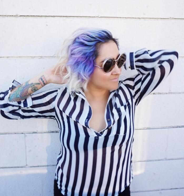 femme avec une coiffure d'ombre inversée de couleur violette, portant des lunettes de soleil et un haut à rayures.