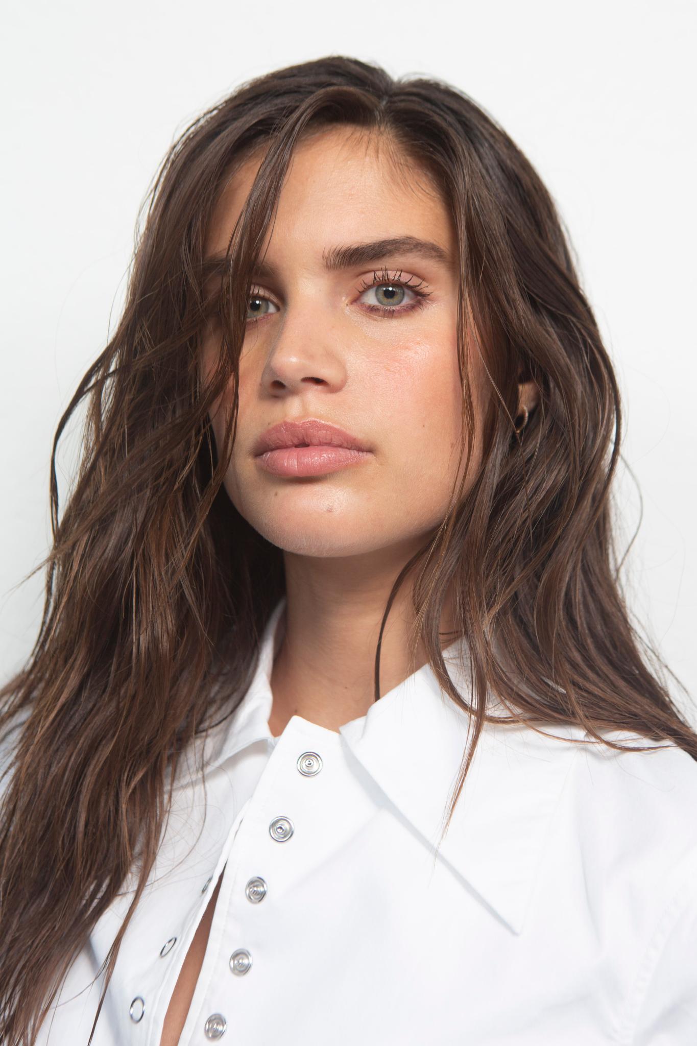 Coiffure d'une invitée de mariage : Vue d'une femme avec de longs cheveux châtain chocolat coiffés en ondulations lâches et portant un haut blanc avec des boutons argentés.