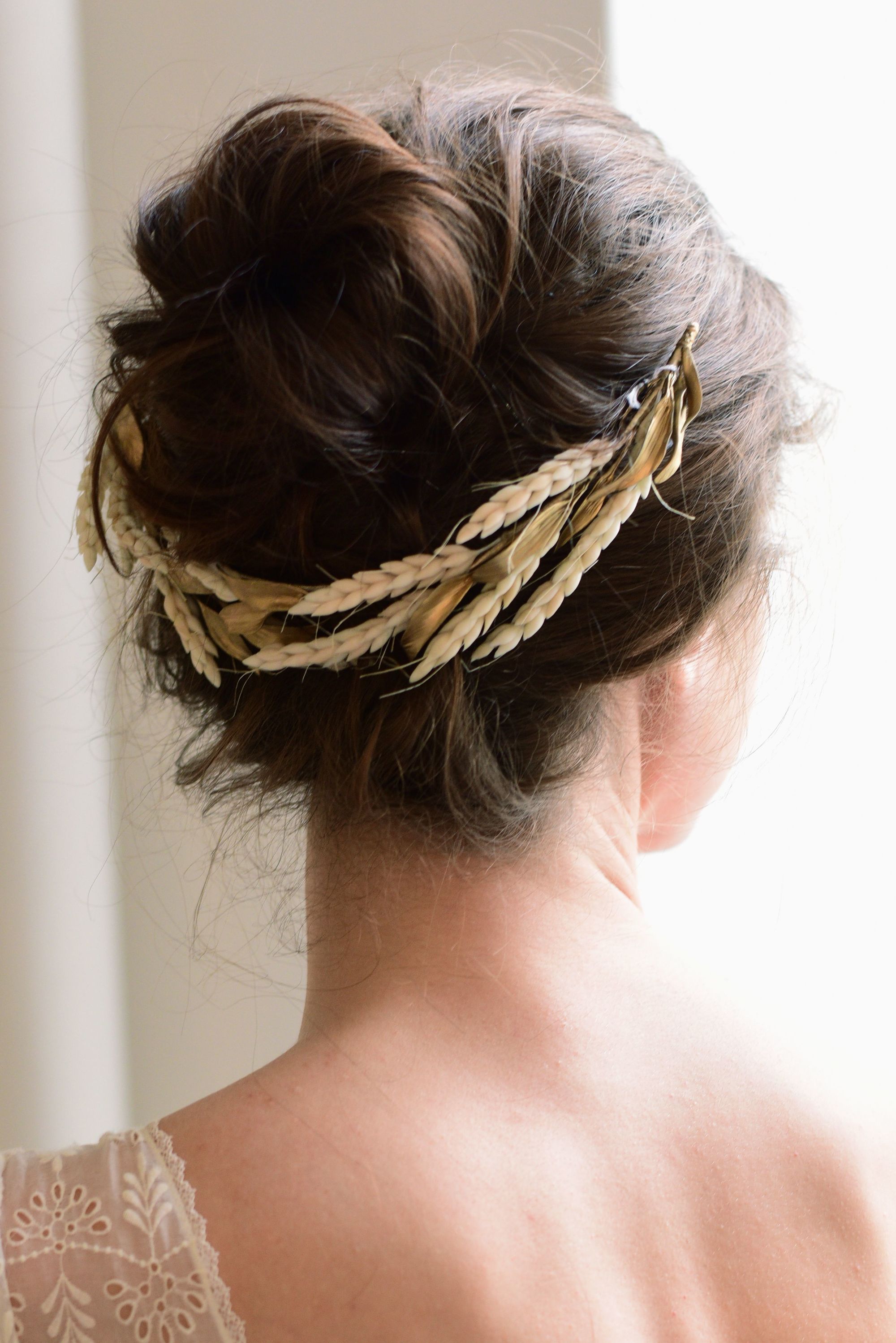 Coiffures d'invités de mariage : Plan arrière d'une femme aux cheveux châtain foncé coiffés en chignon avec un accessoire pour cheveux en forme de fleur dorée, posant dans une pièce éclairée.