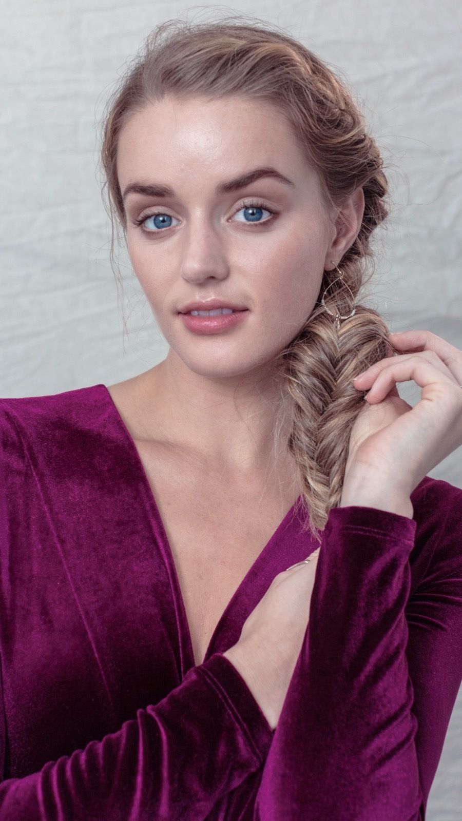 Coiffure d'une invitée de mariage : Vue de côté d'une femme aux cheveux blonds dorés coiffés en tresse en queue de poisson, portant un haut violet et posant dans le studio Poudre de Rose UK.