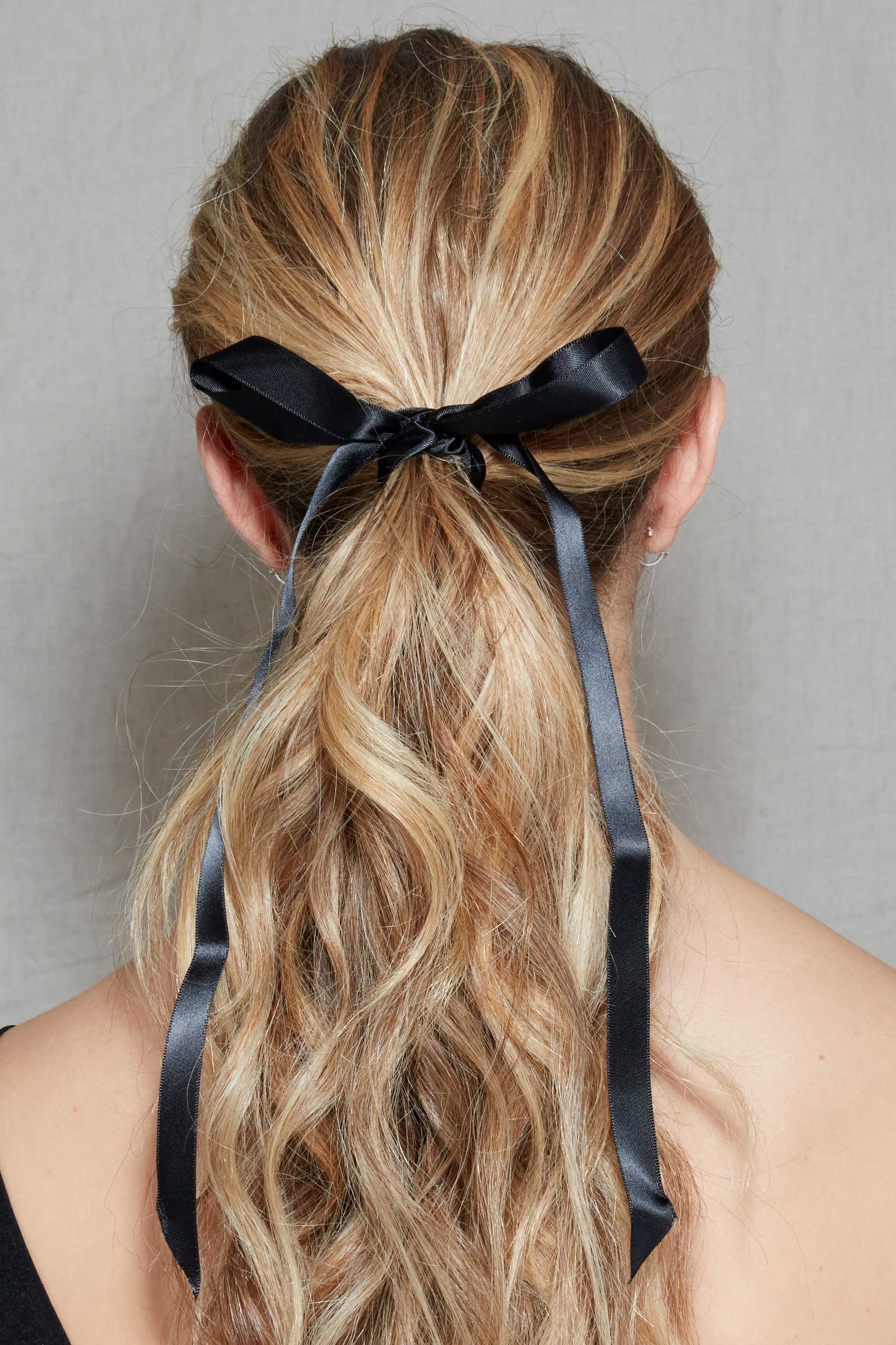 Coiffure d'une invitée de mariage : Plan arrière d'une femme aux longs cheveux blonds sales coiffés en queue de cheval avec un ruban attaché autour, posant dans le studio Poudre de Rose.