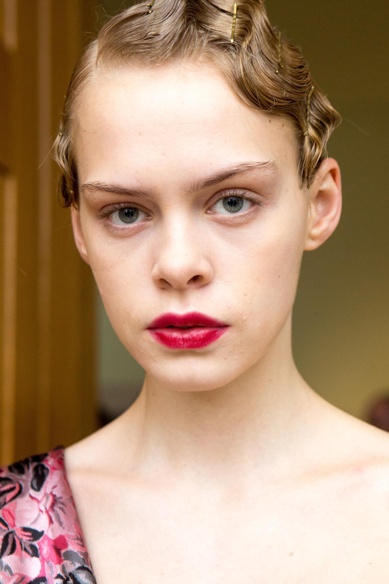 Coiffures pour invités de mariage : Prise de vue d'un mannequin aux cheveux blonds clairs coiffés en ondulations, portant une lèvre cerise et posant dans les coulisses d'un spectacle.