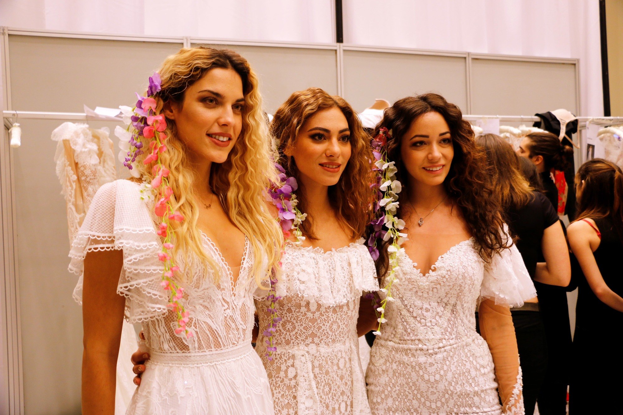 Coiffures d'invitées de mariage : Gros plan de trois femmes aux cheveux de plage bouclés et lâchés, portant du blanc et des fleurs dans les cheveux.