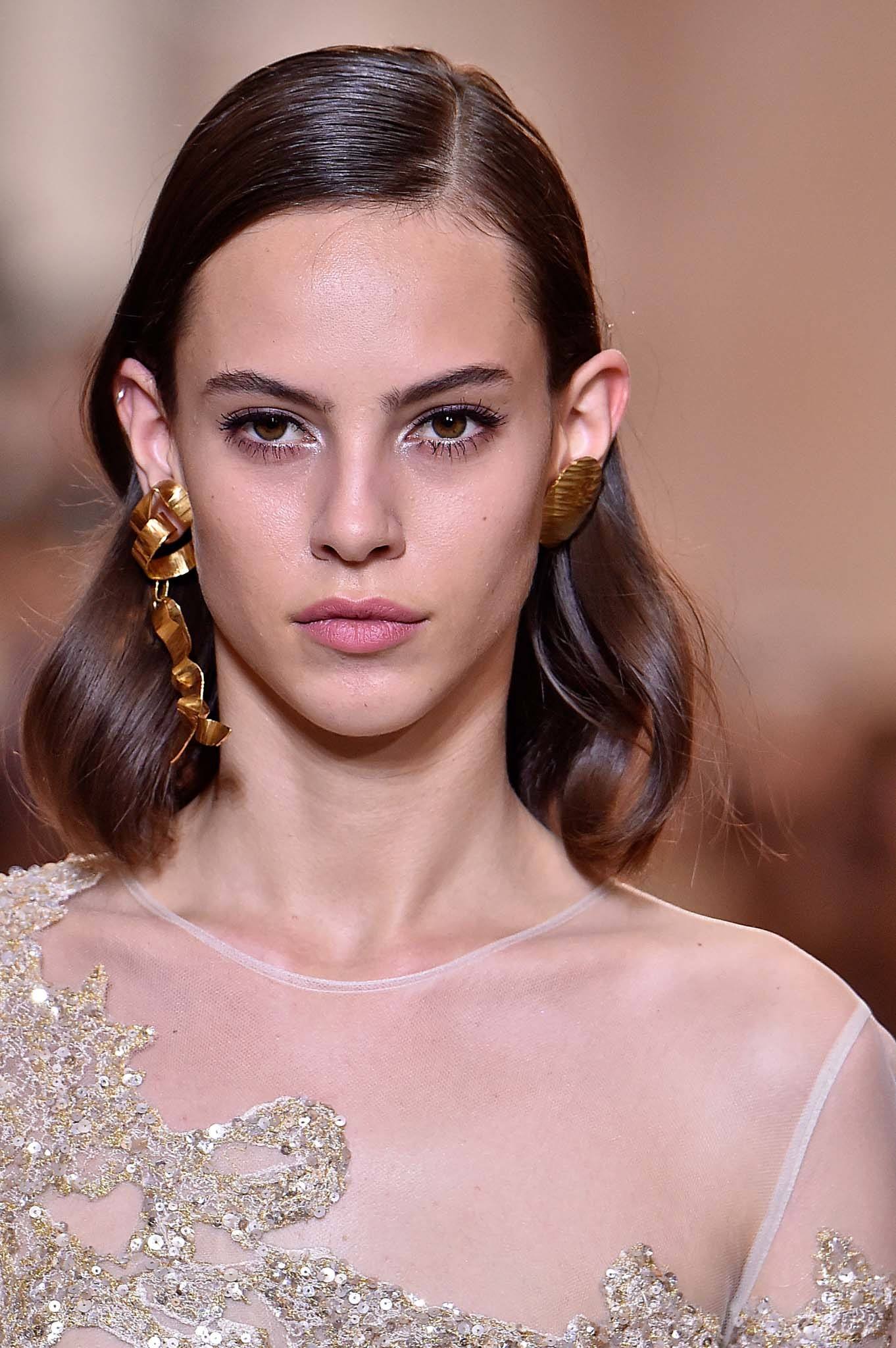 Cheveux d'invités de mariage : Prise de vue d'un mannequin aux vagues brunes moyennes et brillantes sur le podium, portant une robe blanche avec des boucles d'oreilles sur le podium.