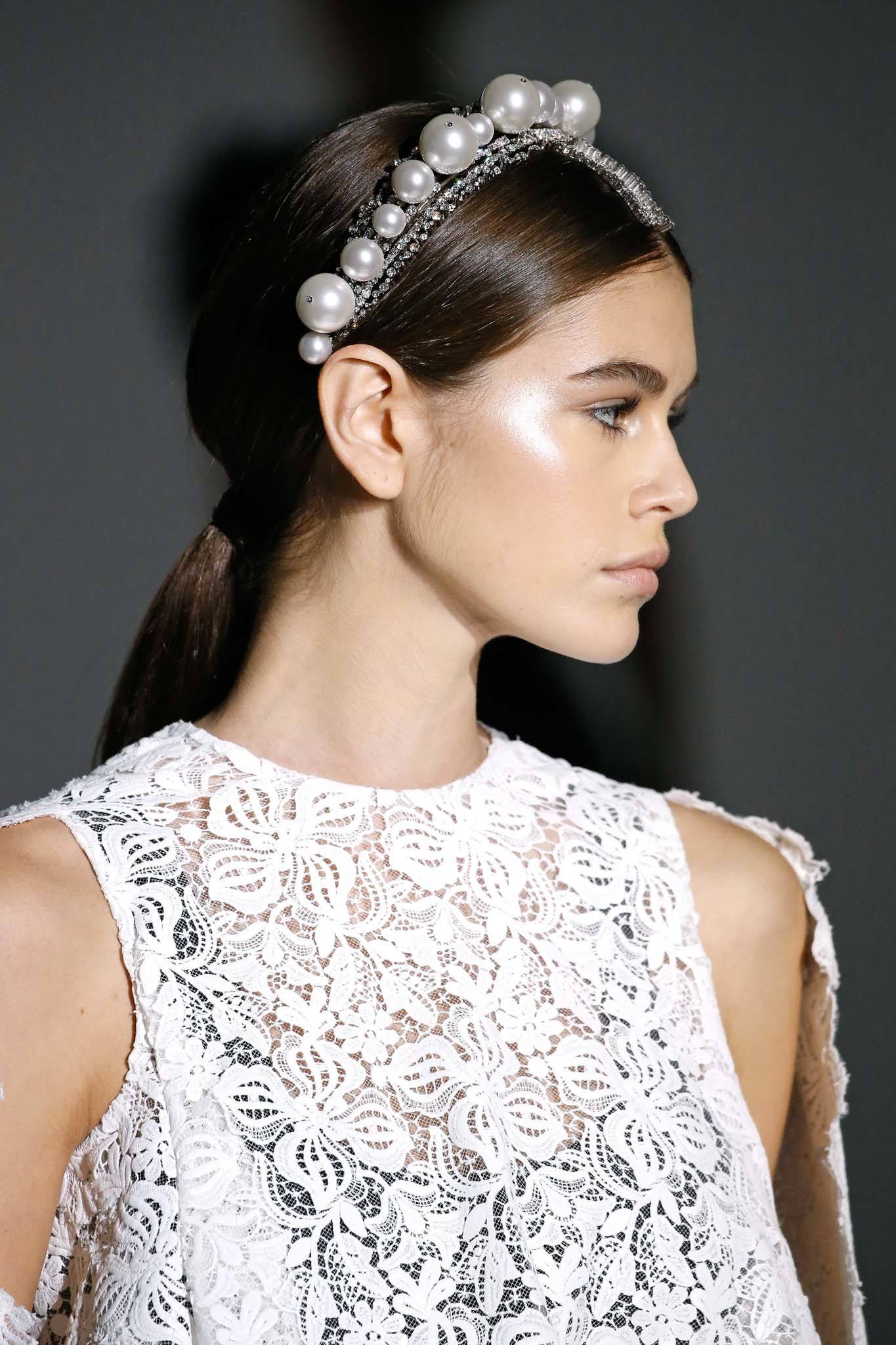 Coiffure d'une invitée de mariage : Prise de vue d'un mannequin aux longs cheveux châtain foncé coiffés en queue de cheval lisse, portant un serre-tête en perles sur le podium.