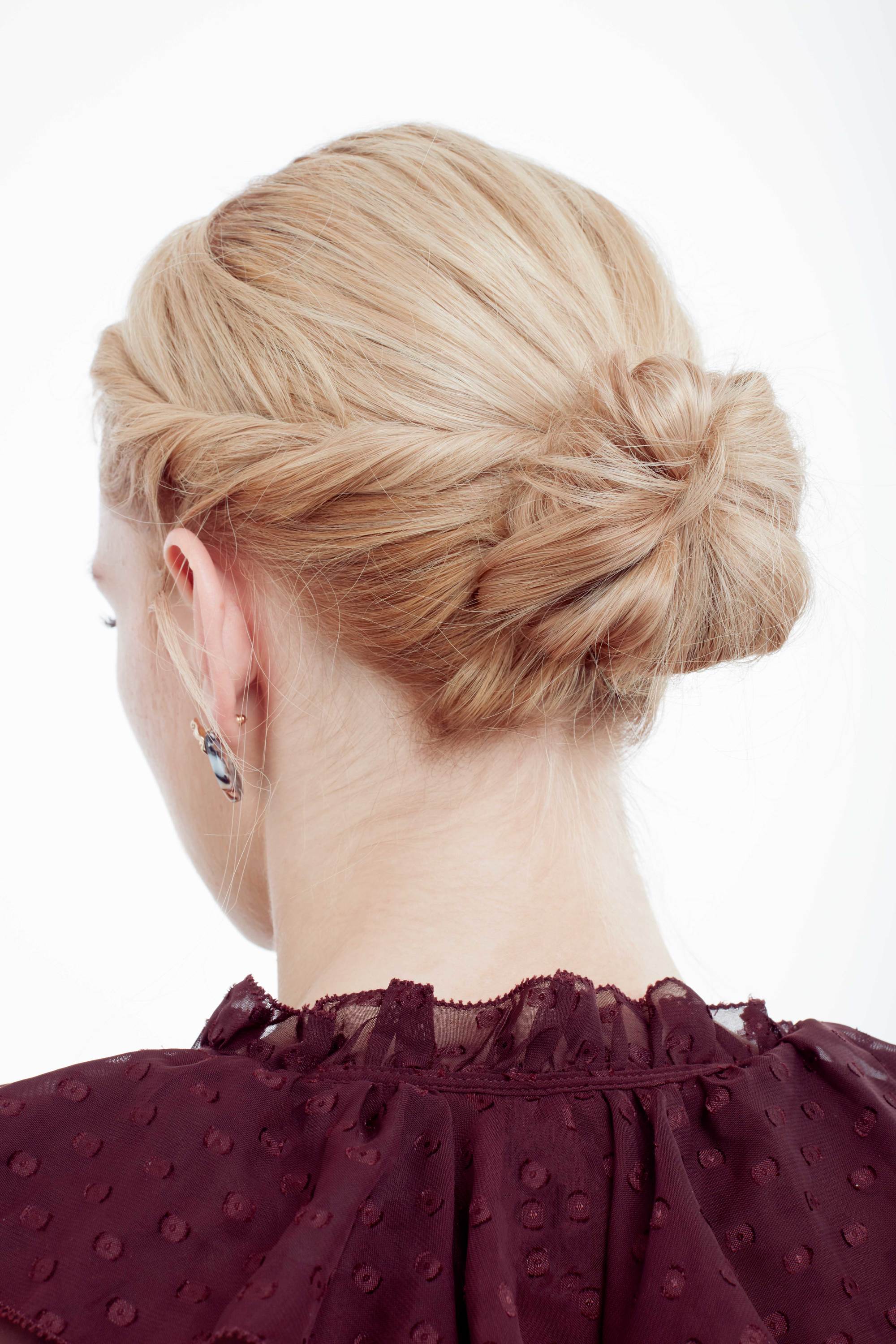 Coiffure d'une invitée de mariage : Plan arrière d'un mannequin aux cheveux blonds dorés coiffés en un chignon torsadé bas, portant un haut violet à volants dans le studio Poudre de Rose UK.