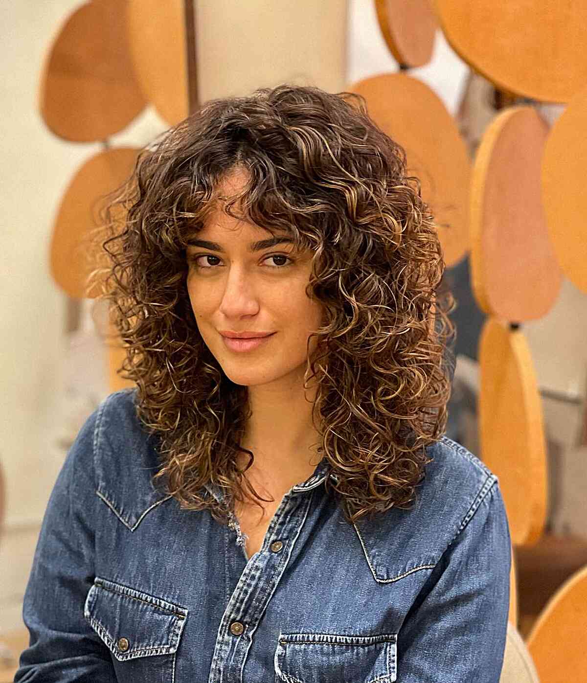 Une femme portant une coupe Cadō mi-longue avec des boucles naturelles et une frange rideau encadrant le visage, idéale pour les textures de cheveux ondulés à bouclés.
