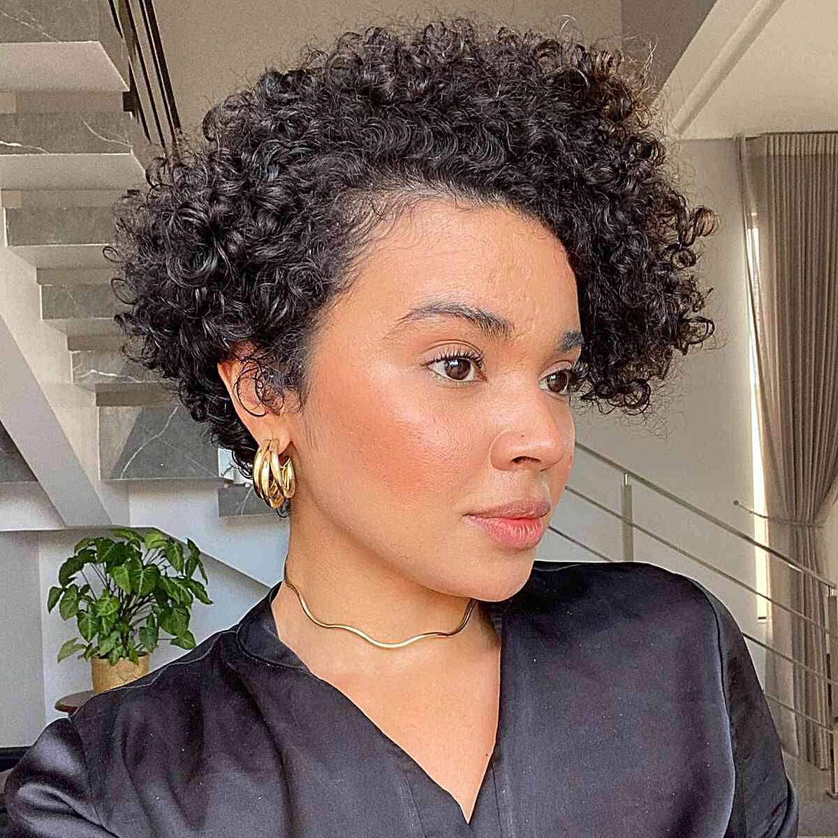 Une femme au visage carré portant une coupe pixie sombre et bouclée, avec des boucles arrondies sur le dessus qui adoucissent sa mâchoire.