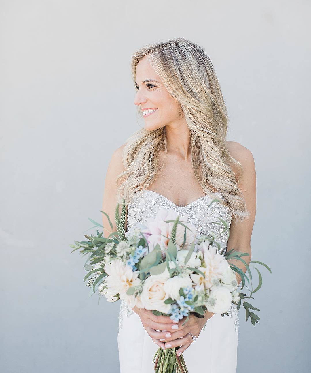 Classic wedding hairstyles: Woman with long bleach blonde hair styled into loose curls, wearing sweetheart wedding dress and holding flowers